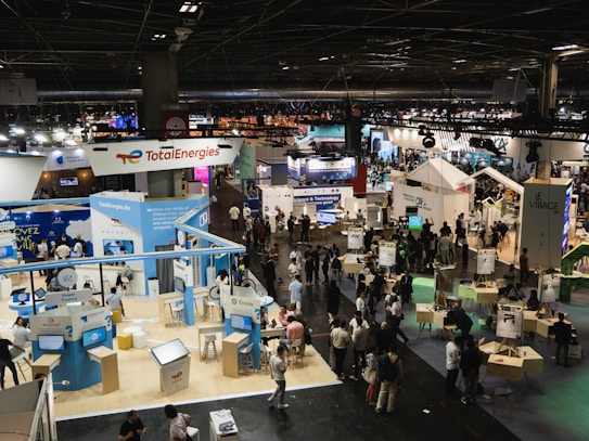 A large, bustling exhibition hall with numerous booths and displays from various companies, some of which have recognizable brand logos. There is a significant number of people walking around, engaging with displays, and interacting with exhibitors. The space is well-lit with overhead lighting, and there are multiple banners and signs throughout the area.