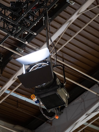 A stage light is suspended from a metal truss in an indoor venue. The light is equipped with barn doors to focus the beam, and there are visible cables attached to it. The background features a ceiling made of wooden beams and metal supports, indicating a warehouse or industrial setting.