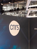 A modern exhibition or event space with industrial lighting and metal structures visible in the background. A large display panel features the acronym 'cnrs' in simple, bold lettering within a white circle. The panel has a dark blue background with subtle geometric patterns.