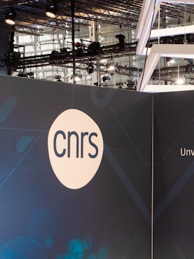 A modern exhibition or event space with industrial lighting and metal structures visible in the background. A large display panel features the acronym 'cnrs' in simple, bold lettering within a white circle. The panel has a dark blue background with subtle geometric patterns.