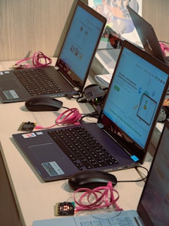 Several laptops with screens displaying a programming interface are arranged on a table. Pink USB cables and mice are connected to the laptops. Small electronic components are also present near the laptops.