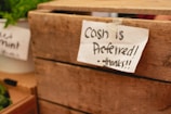 A handwritten note attached to a wooden crate with tape reads 'Cash is preferred! - thanks!!'. The scene appears to be part of a market setting, with fresh greenery visible in the background, likely indicating some produce or herbs.