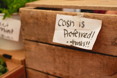 A handwritten note attached to a wooden crate with tape reads 'Cash is preferred! - thanks!!'. The scene appears to be part of a market setting, with fresh greenery visible in the background, likely indicating some produce or herbs.