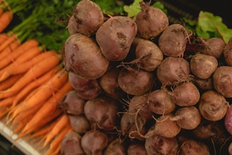 A close-up view of a heap of fresh beets with their roots intact, positioned next to a pile of carrots. The vegetables are placed on what appears to be a market or grocery store display.
