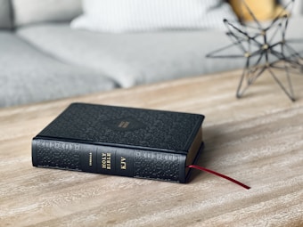 A black leather-bound Bible lies on a wooden table with a red ribbon bookmark. In the background, there's a modern star-shaped metal wire sculpture and a light gray sofa with a white pillow.