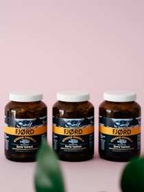 Three brown bottles with white caps are placed side by side on a soft pink surface. The label on the bottles reads 'FJORD STRONG OMEGA-3' and features an image of a fish. The packaging design includes a horizontal orange stripe and some text in a foreign language. Partially blurred green leaves are visible at the bottom of the image.