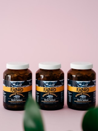 Three brown bottles with white caps are placed side by side on a soft pink surface. The label on the bottles reads 'FJORD STRONG OMEGA-3' and features an image of a fish. The packaging design includes a horizontal orange stripe and some text in a foreign language. Partially blurred green leaves are visible at the bottom of the image.