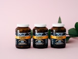 Three amber-colored jars with white caps are aligned on a pink surface. The jars have a label featuring the brand 'FJØRD' with the text 'Strong Omega 3' prominently displayed. A green leaf is partially visible on the right side in the background.