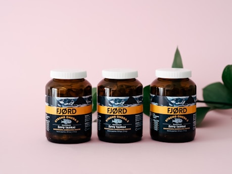 Three amber-colored jars with white caps are aligned on a pink surface. The jars have a label featuring the brand 'FJØRD' with the text 'Strong Omega 3' prominently displayed. A green leaf is partially visible on the right side in the background.