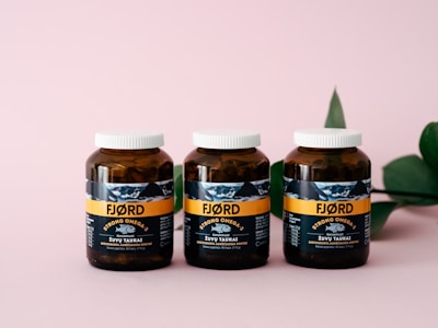 Three amber-colored jars with white caps are aligned on a pink surface. The jars have a label featuring the brand 'FJØRD' with the text 'Strong Omega 3' prominently displayed. A green leaf is partially visible on the right side in the background.