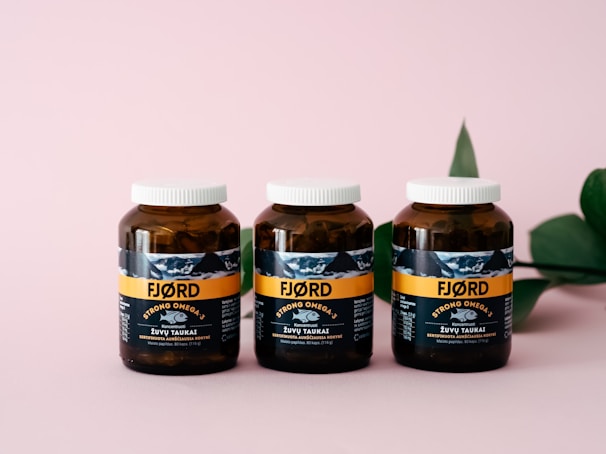 Three amber-colored jars with white caps are aligned on a pink surface. The jars have a label featuring the brand 'FJØRD' with the text 'Strong Omega 3' prominently displayed. A green leaf is partially visible on the right side in the background.