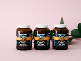 Three amber-colored jars with white caps are aligned on a pink surface. The jars have a label featuring the brand 'FJ&Oslash;RD' with the text 'Strong Omega 3' prominently displayed. A green leaf is partially visible on the right side in the background.