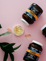 Several jars labeled 'Fjord' containing omega-3 capsules are arranged on a pink surface. The jars are dark with white caps, while the labels are predominantly yellow and black. Some capsules are placed in a small dish and scattered around. Green leaves serve as an accent in the composition.