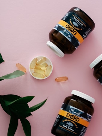 Several jars labeled 'Fjord' containing omega-3 capsules are arranged on a pink surface. The jars are dark with white caps, while the labels are predominantly yellow and black. Some capsules are placed in a small dish and scattered around. Green leaves serve as an accent in the composition.