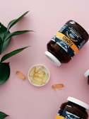 A flat lay composition featuring two large brown supplement jars with white caps lying on a light pink background. The jars are labeled with a black, white, and gold design. In between the jars, a small, white dish contains several yellowish oval capsules. There are also three standalone capsules scattered near the dish. A green leafy plant is positioned in the top left corner, adding a touch of natural greenery to the scene.