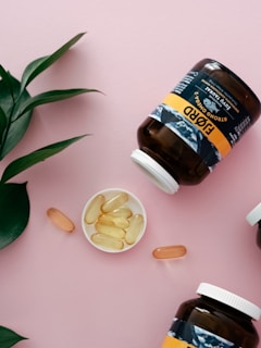 A flat lay composition featuring two large brown supplement jars with white caps lying on a light pink background. The jars are labeled with a black, white, and gold design. In between the jars, a small, white dish contains several yellowish oval capsules. There are also three standalone capsules scattered near the dish. A green leafy plant is positioned in the top left corner, adding a touch of natural greenery to the scene.