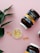 A flat lay composition featuring two large brown supplement jars with white caps lying on a light pink background. The jars are labeled with a black, white, and gold design. In between the jars, a small, white dish contains several yellowish oval capsules. There are also three standalone capsules scattered near the dish. A green leafy plant is positioned in the top left corner, adding a touch of natural greenery to the scene.