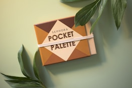 A makeup pocket palette with a geometric design, featuring the brand name written on it, surrounded by green leaves on a soft green background.