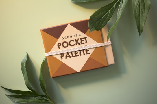 A makeup pocket palette with a geometric design, featuring the brand name written on it, surrounded by green leaves on a soft green background.