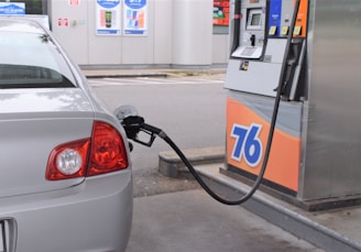 A silver car is parked at a gas station with a fuel nozzle inserted into its fuel tank. The gas pump shows the number 76 and various buttons and displays for fuel options. In the background, a station wall with advertisements and price displays can be seen.
