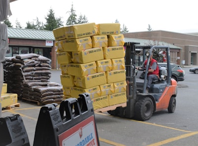 A forklift operator is moving a large stack of yellow bags in a parking lot, with additional stacks of bags in the background. The forklift is orange and black, with a person in a red shirt driving it. Nearby, there are signs and parked cars, and the scene takes place outside a commercial building with a green metal roof.