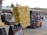 A forklift operator is moving a large stack of yellow bags in a parking lot, with additional stacks of bags in the background. The forklift is orange and black, with a person in a red shirt driving it. Nearby, there are signs and parked cars, and the scene takes place outside a commercial building with a green metal roof.
