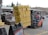 A forklift operator is moving a large stack of yellow bags in a parking lot, with additional stacks of bags in the background. The forklift is orange and black, with a person in a red shirt driving it. Nearby, there are signs and parked cars, and the scene takes place outside a commercial building with a green metal roof.