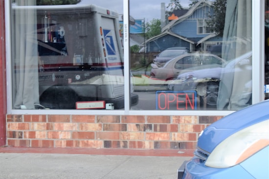 A storefront window displaying a neon sign that reads 'OPEN'. A vehicle, possibly a postal truck, is reflected in the glass. The scene includes the reflection of parked cars and part of a blue vehicle in front, with a residential house visible in the background.
