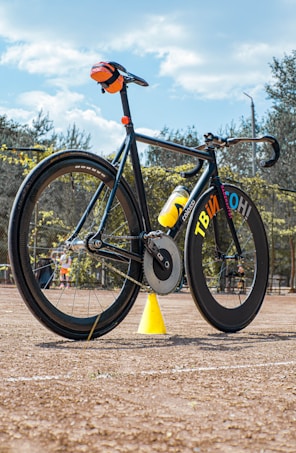 A sleek, modern bicycle is positioned on a sandy terrain, accentuated by its black and yellow color scheme. The bike features large, aerodynamic wheels, a water bottle mounted on the frame, and a small orange bag attached to the seat. In the background, there are trees and a blue sky with a few scattered clouds overhead. A bright yellow cone stands near the bicycle, adding a striking contrast.