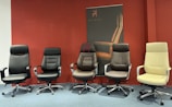 Five modern office chairs of various styles and colors are arranged in a row against a wall with a dark red background. Behind them is a promotional poster with the brand 'MyChair'. The chairs have different designs, armrests, and backrest styles, showcasing different ergonomic features.