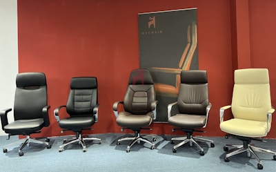 Five modern office chairs of various styles and colors are arranged in a row against a wall with a dark red background. Behind them is a promotional poster with the brand 'MyChair'. The chairs have different designs, armrests, and backrest styles, showcasing different ergonomic features.