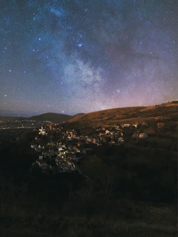 A serene village nestled in rolling hills under a starry sky, inviting quiet reflection.