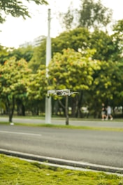 A small drone hovers in the air against a backdrop of a lush green park with trees. A road runs alongside the park, with a blurred pedestrian in the background. The setting appears peaceful and natural.