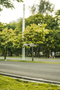A small drone hovers in the air against a backdrop of a lush green park with trees. A road runs alongside the park, with a blurred pedestrian in the background. The setting appears peaceful and natural.