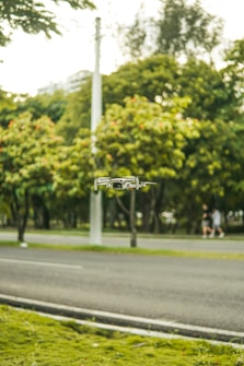 A small drone hovers in the air against a backdrop of a lush green park with trees. A road runs alongside the park, with a blurred pedestrian in the background. The setting appears peaceful and natural.
