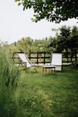 A serene garden scene with two white deck chairs positioned on a lush green lawn. A small wooden table sits between the chairs. The background features a wooden fence and a variety of green leafy trees and shrubs, creating a peaceful outdoor setting.