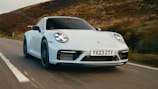 A sleek white sports car speeding down a rural road with blurred vegetation in the background, suggesting motion. The car displays a license plate with the letters and numbers YX23 ZTY.