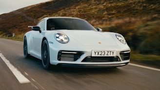 A sleek white sports car speeding down a rural road with blurred vegetation in the background, suggesting motion. The car displays a license plate with the letters and numbers YX23 ZTY.