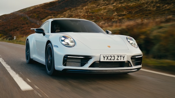 A sleek white sports car speeding down a rural road with blurred vegetation in the background, suggesting motion. The car displays a license plate with the letters and numbers YX23 ZTY.