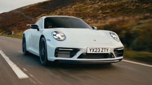 A sleek white sports car speeding down a rural road with blurred vegetation in the background, suggesting motion. The car displays a license plate with the letters and numbers YX23 ZTY.