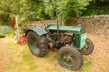 An old green tractor is parked on grassy ground near a stone wall. The tractor has large black tires and a slightly rusted exterior. Behind it, there is a piece of red and yellow agricultural equipment. The background includes lush green trees and bushes, creating a serene rural setting. A couple of blue folding chairs are visible to the left of the tractor.