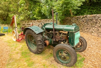 An old green tractor is parked on grassy ground near a stone wall. The tractor has large black tires and a slightly rusted exterior. Behind it, there is a piece of red and yellow agricultural equipment. The background includes lush green trees and bushes, creating a serene rural setting. A couple of blue folding chairs are visible to the left of the tractor.
