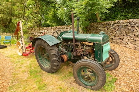 An old green tractor is parked on grassy ground near a stone wall. The tractor has large black tires and a slightly rusted exterior. Behind it, there is a piece of red and yellow agricultural equipment. The background includes lush green trees and bushes, creating a serene rural setting. A couple of blue folding chairs are visible to the left of the tractor.