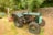 An old green tractor is parked on grassy ground near a stone wall. The tractor has large black tires and a slightly rusted exterior. Behind it, there is a piece of red and yellow agricultural equipment. The background includes lush green trees and bushes, creating a serene rural setting. A couple of blue folding chairs are visible to the left of the tractor.