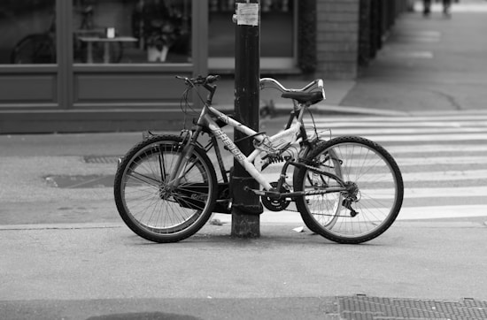 A bicycle is leaning against a metal post on a city sidewalk. The bike appears slightly worn and is parked near a zebra crossing. In the background, there is a shop with large windows, and further down the street, a few more buildings are visible.