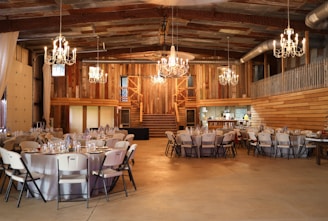 A rustic barn interior is set up for an event with round tables covered in white tablecloths and surrounded by folding chairs. Elaborate chandeliers hang from the ceiling, casting warm light across the wooden walls and exposed beams. There is a staircase in the background leading to a second level. The venue gives off a cozy and inviting ambiance.