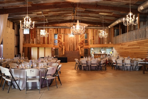 A rustic barn interior is set up for an event with round tables covered in white tablecloths and surrounded by folding chairs. Elaborate chandeliers hang from the ceiling, casting warm light across the wooden walls and exposed beams. There is a staircase in the background leading to a second level. The venue gives off a cozy and inviting ambiance.