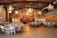 A rustic barn interior is set up for an event with round tables covered in white tablecloths and surrounded by folding chairs. Elaborate chandeliers hang from the ceiling, casting warm light across the wooden walls and exposed beams. There is a staircase in the background leading to a second level. The venue gives off a cozy and inviting ambiance.