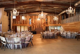 A rustic barn interior is set up for an event with round tables covered in white tablecloths and surrounded by folding chairs. Elaborate chandeliers hang from the ceiling, casting warm light across the wooden walls and exposed beams. There is a staircase in the background leading to a second level. The venue gives off a cozy and inviting ambiance.