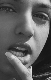 A close-up black and white portrait of a person's face focusing on the eyes, nose, and mouth. The person has a finger touching their lower lip, showcasing detailed facial features and texture.